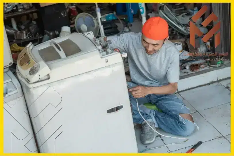 شخص يرتدي قميصًا رمادي اللون وقبعة برتقالية يقوم بإصلاح غسالة ملابس على الأرضية المبلطة ببراعة، مستعرضًا مهاراته في صيانة غسالات مكة. تنتشر الأدوات والأجزاء المختلفة في كل مكان، تجسد تفانيهم في حرفتهم.