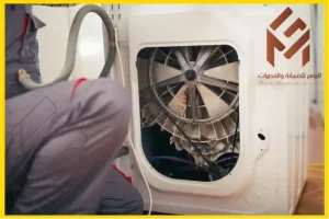 فني يقوم بإصلاح صيانة غسالة دايو بمكة، يظهر الجزء الداخلي وتروس المحرك، مع شعار شركة "الماهر للصيانة والخدمات" في الزاوية.