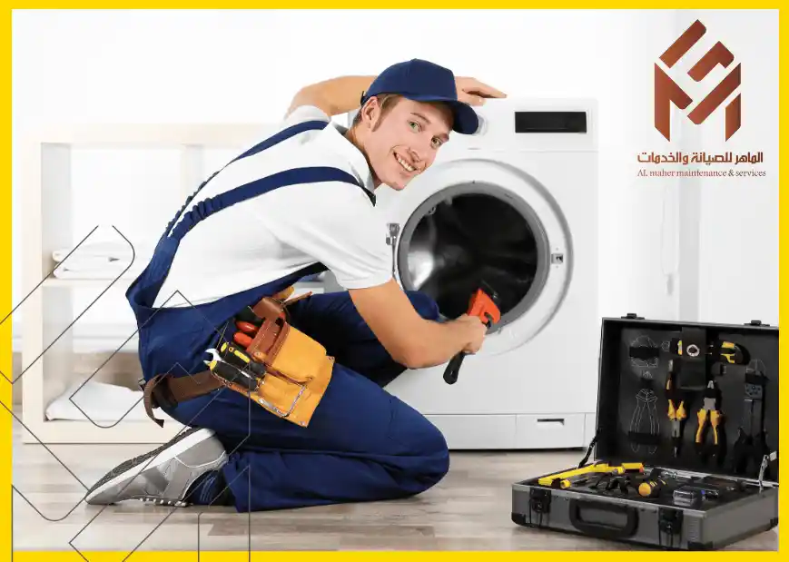 فني صيانة يرتدي زي عمل أزرق وقبعة، يقوم بفحص أو إصلاح غسالة أمامية، مع حقيبة أدوات مفتوحة بجانبه، وشعار شركة على الحائط مكتوب عليه "الماهر للصيانة والخدمات".  صيانة غسالة ال جي بمكة المكرمة.