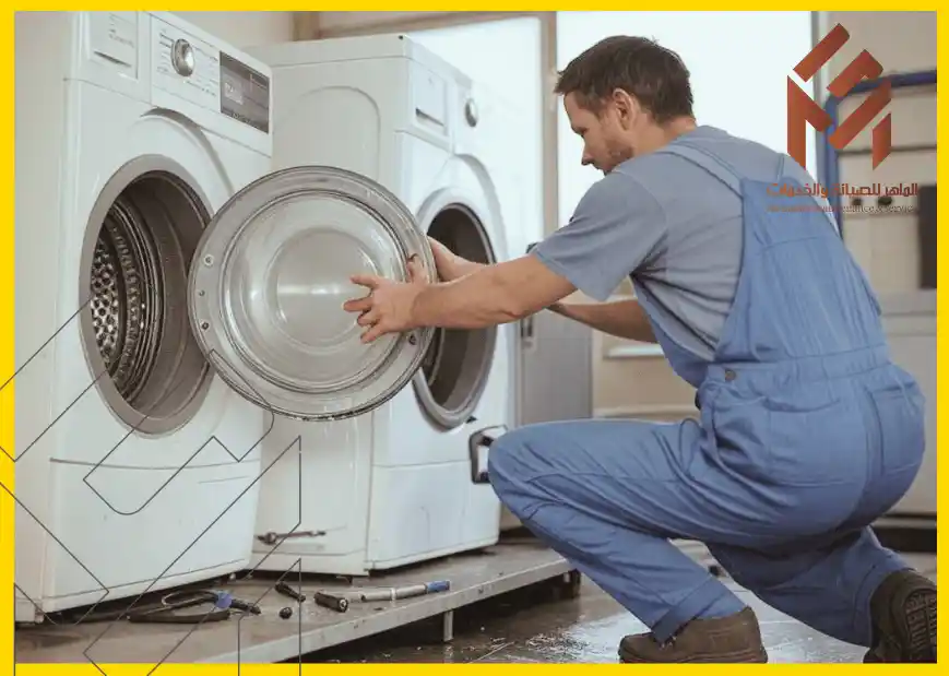 مهندس غسالات اتوماتيك بمكة يرتدي زي عمل أزرق يعمل على إصلاح غسالة ملابس بفتحة أمامية في غرفة غسيل.