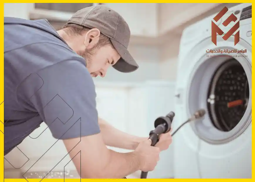 رجل يرتدي قبعة وقميص أزرق يقوم بفحص أو تركيب غسالة في مطبخ، مع شعار شركة في الزاوية العليا اليمنى للصورة.حلول في غسالات الملابس
