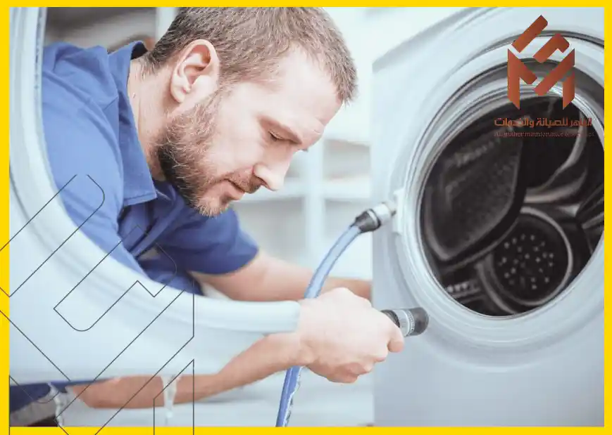 رجل يرتدي زي عمل أزرق يقوم بفحص أو إصلاح غسالة، بينما يمسك بأنبوب موصل بها.فني صيانة غسالات ملابس في مكة