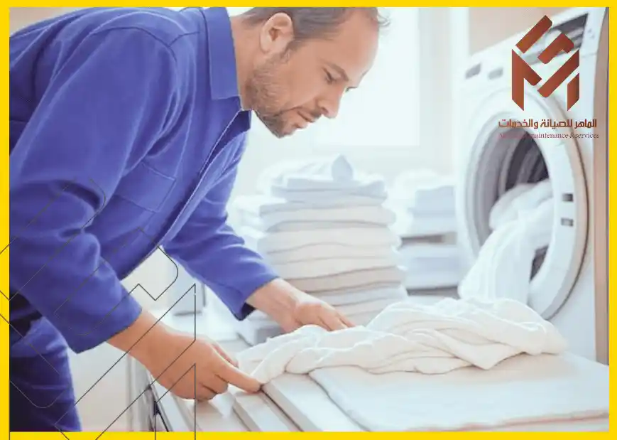 رجل يرتدي زي عمل أزرق يقوم بترتيب المناشف البيضاء بجانب غسالة في غرفة غسيل.المشكلات الشائعة في غسالات الملابس
