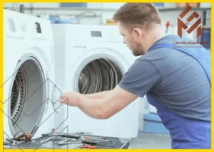 رجل يرتدي زي عمل أزرق يقوم بصيانة غسالة بيضاء اللون، مع مجموعة متنوعة من الأدوات الموضوعة على الطاولة بجانبه وشعار الشركة في الزاوية اليمنى العليا.مشكلات غسالات الملابس