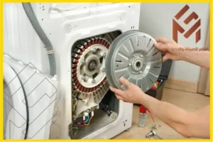 شخص يقوم بفك أو تركيب دائرة محرك غسالة أوتوماتيكية بيضاء، ويظهر جانبها الداخلي والمكونات الكهربائية. توجد أدوات على الأرض وشعار باللغة العربية في الأعلى على اليمين.مزايا وعيوب محرك الدفع المباشر في غسالة الملابس